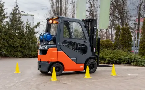 Pomarańczowo-szary wózek widłowy Toyota z butlą gazową, otoczony żółtymi pachołkami. Zastanawiasz się, ile trwa kurs na wózek widłowy?