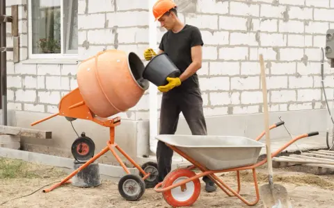 Budowlaniec wsypuje cement do betoniarki 100l. Obok stoi taczka i łopata.