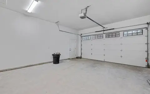 Nowy garaż z płyt betonowych projekt. Biała brama segmentowa, drzwi, czarny kosz na śmieci i sufitowe oświetlenie.