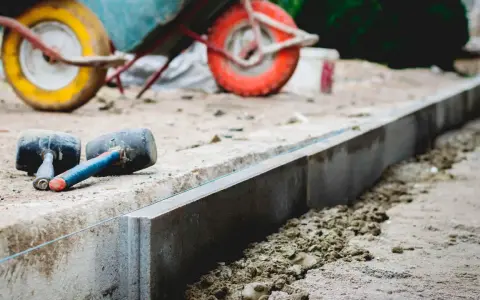 Dwa gumowe młotki leżą obok świeżo ułożonych krawężników. W tle widać taczkę, sugerującą prace budowlane i potrzebę odpowiedniej podsypki pod krawężniki proporcje.