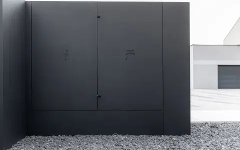 Nowoczesna, czarna skrzynka elektryczna w ogrodzeniu, z dyskretnymi symbolami błyskawicy i kranu.