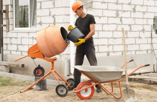 Budowlaniec wsypuje cement do betoniarki 100l. Obok stoi taczka i łopata.