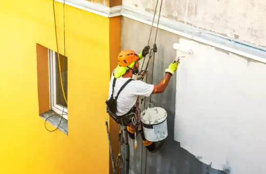 Pracownik na wysokości maluje elewację. Sprawdź, jaka jest malowanie elewacji cena i jak odnowić budynek.
