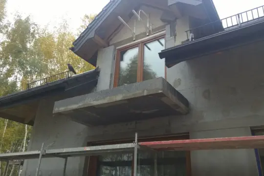 Budynek w trakcie ocieplania elewacji. Widać rusztowania, szare ściany i fragment dachu z ptakiem.