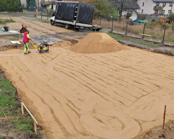 Pracownik zagęszcza piasek zagęszczarką. To kluczowy etap wymiany gruntu pod płytę fundamentową.