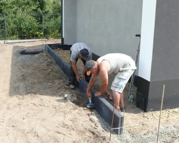 Dwóch mężczyzn układa suchy beton pod obrzeża wokół domu.