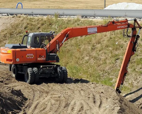 Pomarańczowa koparka Hitachi Zaxis 160W pracuje na budowie. Operator z uprawnieniami na minikoparkę wykonuje prace ziemne.