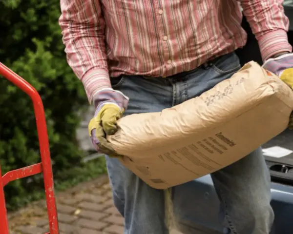 Mężczyzna w rękawicach niesie worek cementu. Zastanawia się, ile piasku na 25 kg cementu będzie potrzebne do budowy.