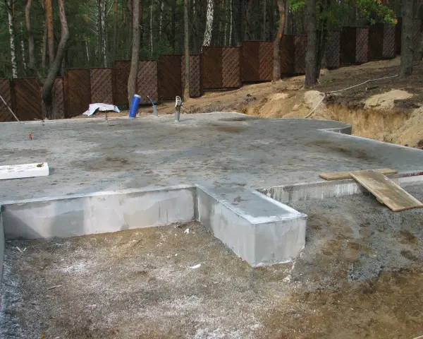 Solidna płyta fundamentowa, gotowa na budowę domu. Wokół las i drewniany płot.
