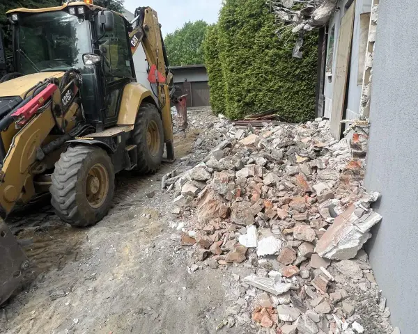 Koparka pracuje przy rozbiórce domu. Gruz i cegły leżą na ziemi, szacowany koszt rozbiórki domu jest wysoki.