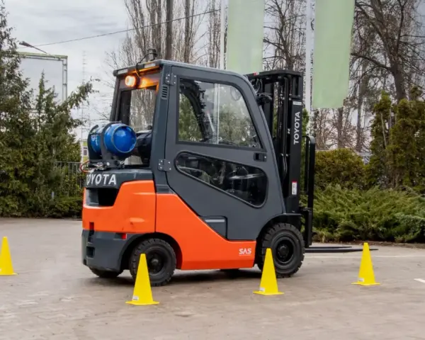 Pomarańczowo-szary wózek widłowy Toyota z butlą gazową, otoczony żółtymi pachołkami. Zastanawiasz się, ile trwa kurs na wózek widłowy?