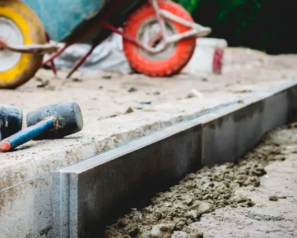 Dwa gumowe młotki leżą obok świeżo ułożonych krawężników. W tle widać taczkę, sugerującą prace budowlane i potrzebę odpowiedniej podsypki pod krawężniki proporcje.