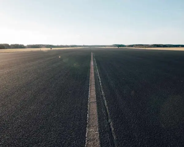 Asfaltowa droga z linią pośrodku, otoczona polami i lasem.