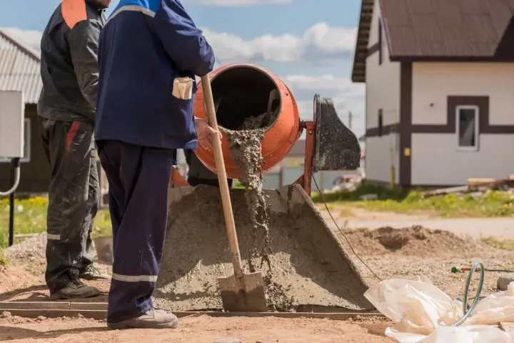 Dwóch budowlańców miesza beton w betoniarce. Widać, ile żwiru na worek cementu potrzeba do uzyskania odpowiedniej konsystencji.