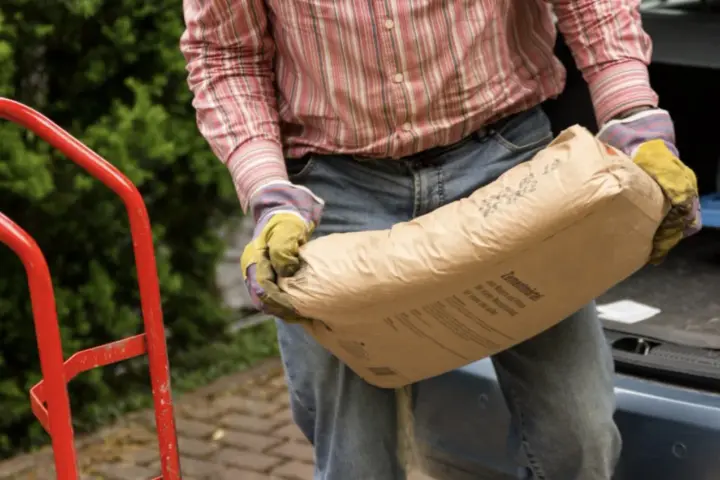 Mężczyzna w rękawicach niesie worek cementu. Zastanawia się, ile piasku na 25 kg cementu będzie potrzebne do budowy.