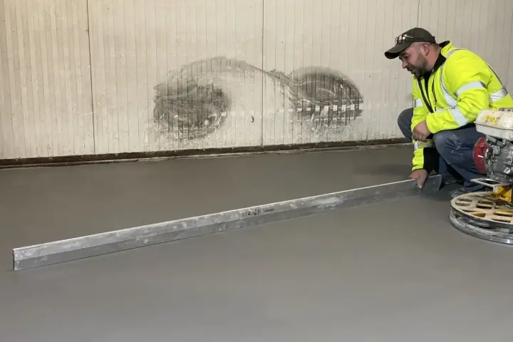 Pracownik w kamizelce odblaskowej wykonuje poziomowanie wylewki betonowej za pomocą łaty.