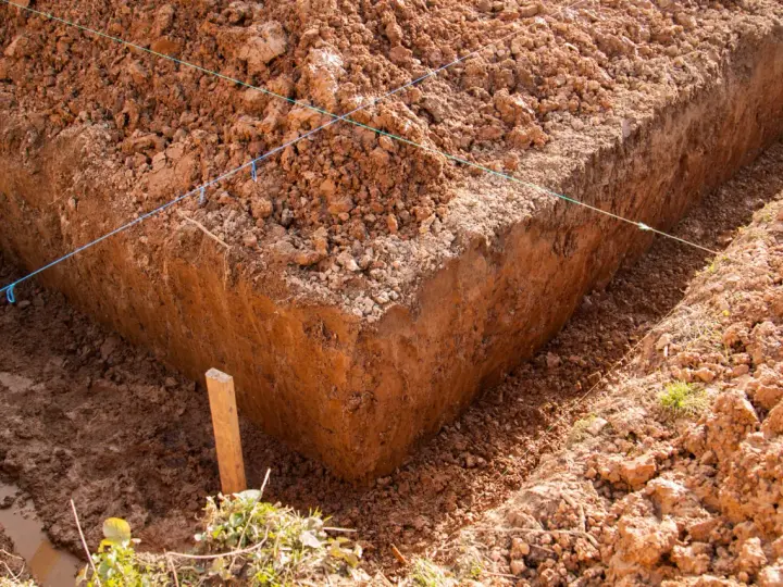 Głęboki wykop pod fundament, z widocznymi nićmi wyznaczającymi linie budowy. Widać błotniste dno i usypaną ziemię.