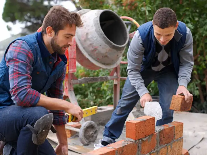 Dwóch budowlańców układa cegły. Jeden sprawdza poziom, drugi kładzie cegłę. W tle betoniarka, która pomoże ustalić, ile cementu na kubik betonu.
