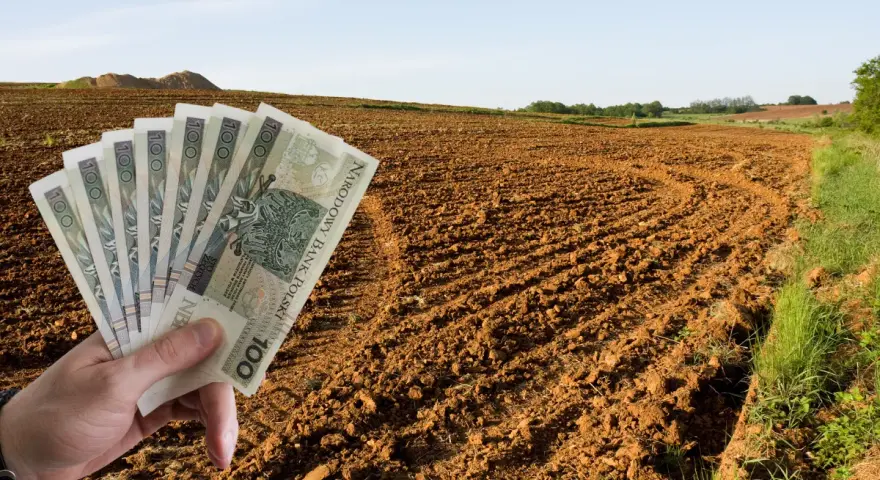 Dłoń trzyma banknoty 100 zł na tle zaoranego pola. Zastanawiasz się, ile kosztuje ar ziemi rolnej?
