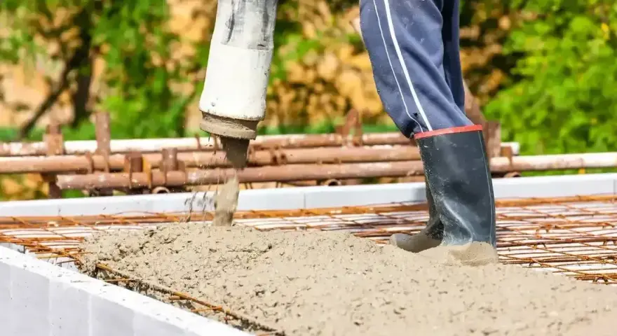 Beton wylewany do szalunków fundamentowych. Pracownik w gumowcach nadzoruje proces.