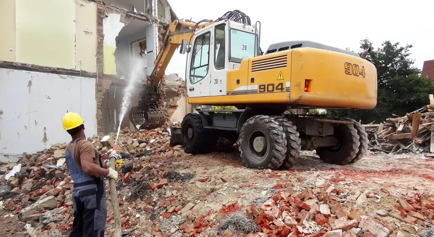 Robot budowlany pracuje przy wyburzaniu budynku. Robotnik z węża zrasza gruz, by ograniczyć pylenie. Szacowany koszt wyburzenia domu 100m2.