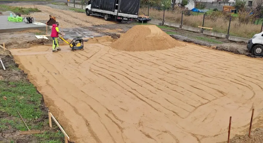Pracownik zagęszcza piasek zagęszczarką. To kluczowy etap wymiany gruntu pod płytę fundamentową.