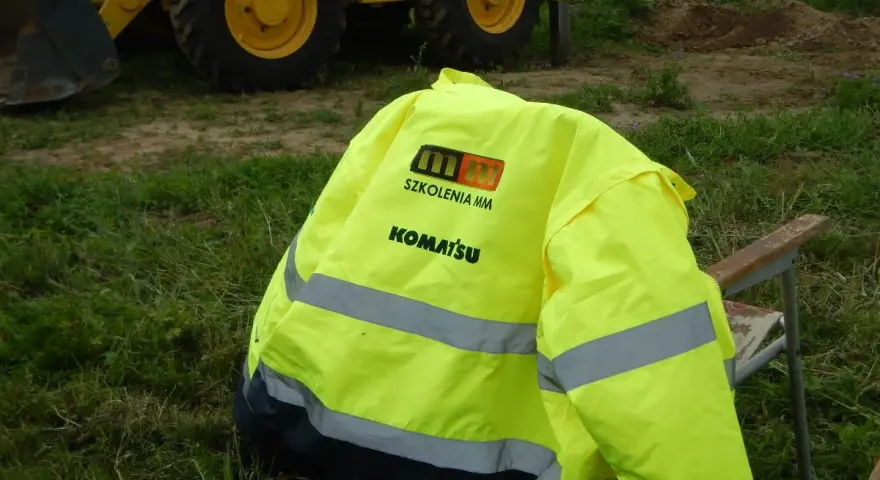 Żółta kamizelka z logo Komatsu i napisem "Szkolenia MM" leży na krześle. To przygotowanie do egzaminu na ładowarkę, gdzie pytania i odpowiedzi są kluczowe.