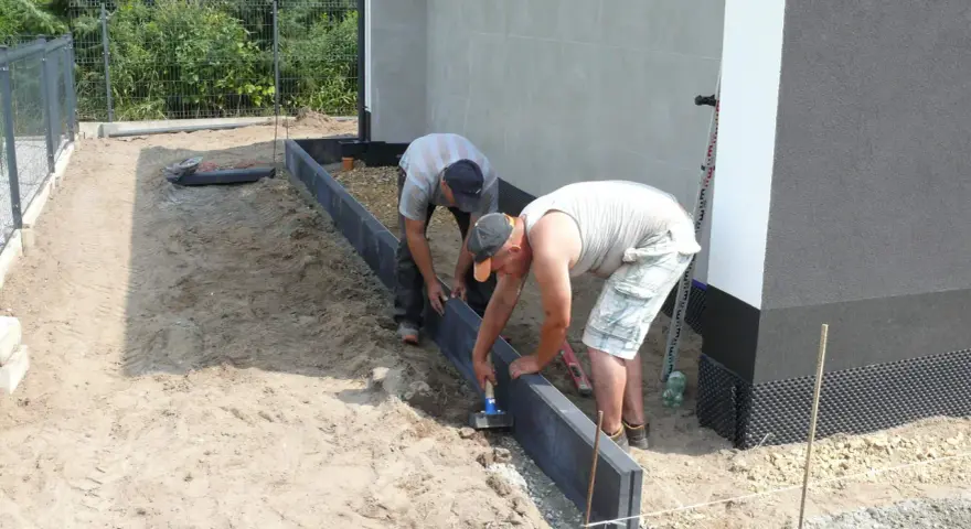 Dwóch mężczyzn układa suchy beton pod obrzeża wokół domu.