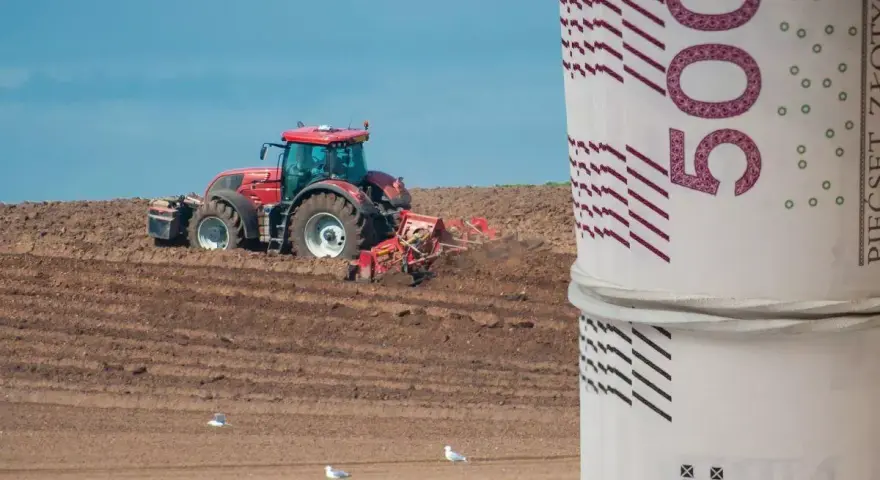 Czerwony traktor orze pole, a na pierwszym planie zwinięte banknoty 500 zł. Zastanawiasz się, ile kosztuje hektar ziemi budowlanej?