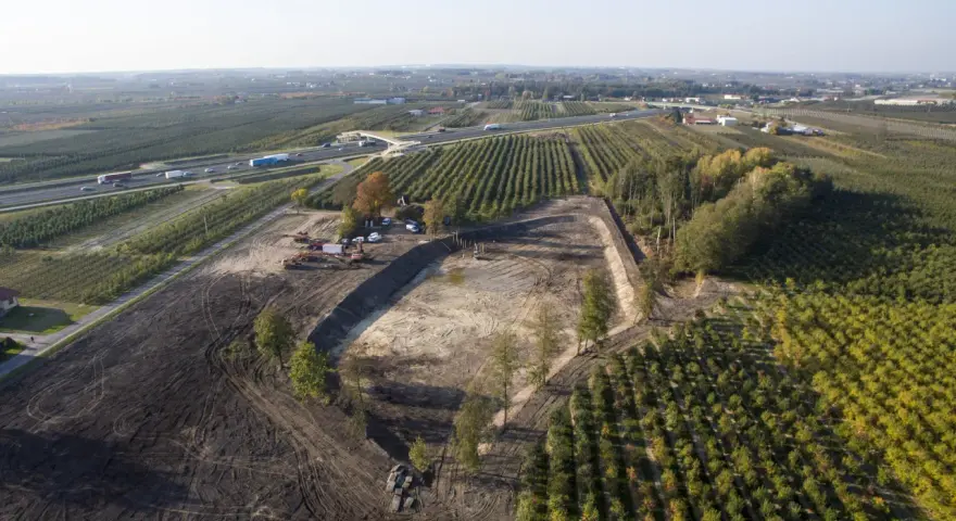 Trwa budowa stawu rybnego. Widać koparki, maszyny budowlane i przygotowany teren pod zbiornik wodny, otoczony sadem i autostradą.