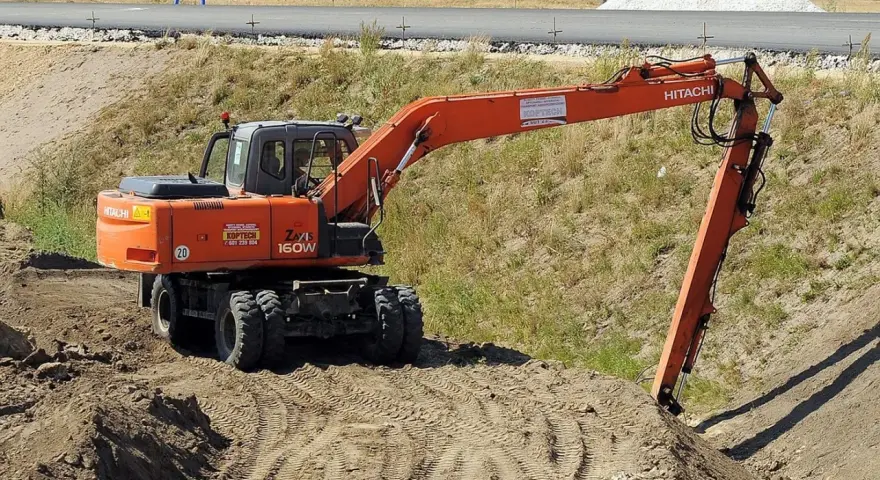 Pomarańczowa koparka Hitachi Zaxis 160W pracuje na budowie. Operator z uprawnieniami na minikoparkę wykonuje prace ziemne.