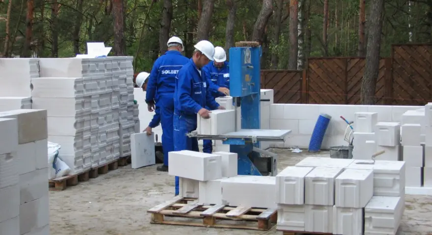 Budowa trwa! Pracownicy w niebieskich kombinezonach i białych kaskach układają bloczki ytong. Na paletach widać, że ytong 12 to spora ilość materiału budowlanego.
