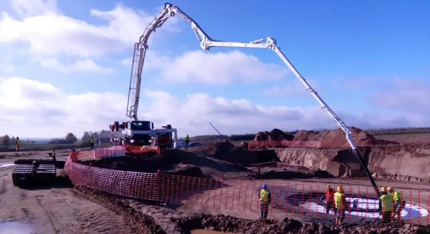 Ogromna pompa do betonu pracuje na budowie, dostarczając materiał do wykopu. Pracownicy w kamizelkach obserwują proces.