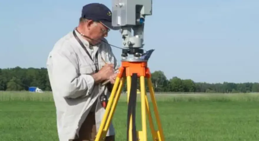 Geodeta precyzyjnie ustala granice działki, używając nowoczesnego sprzętu pomiarowego na tle zielonego pola.