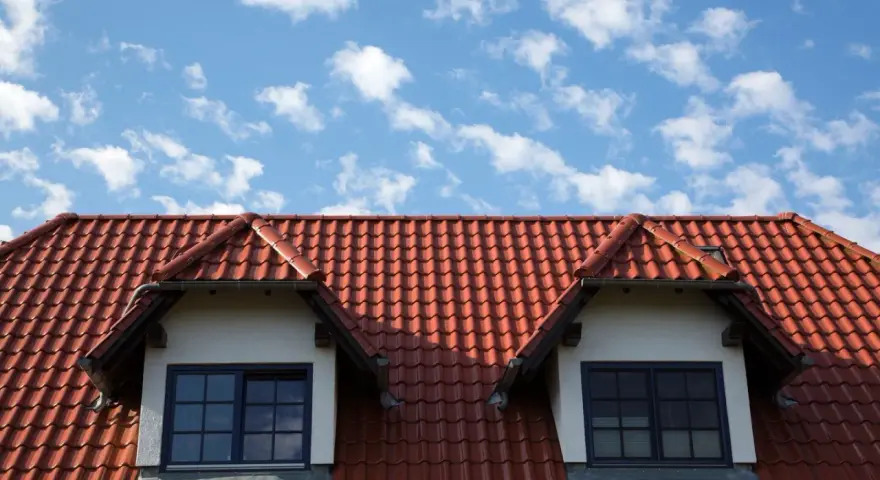Czerwony dach z dachówki ceramicznej, z dwoma lukarnami i niebieskim niebem w chmurach.