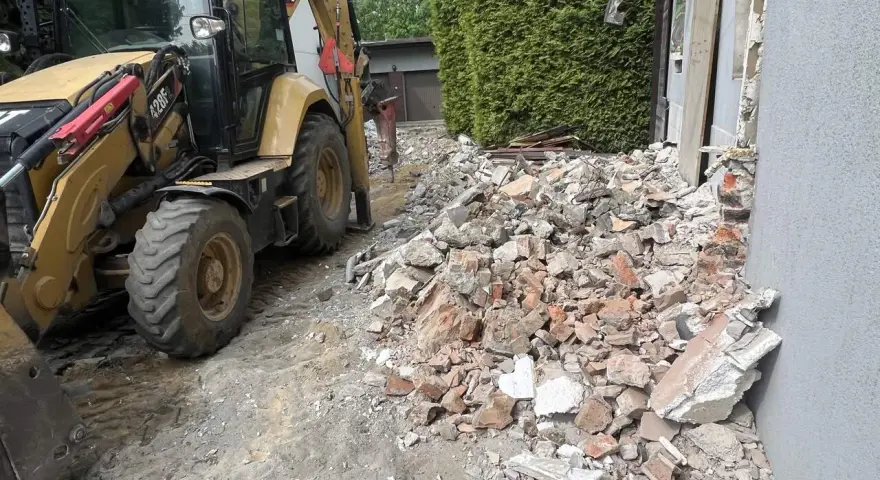Żółta koparko-ładowarka pracuje przy wyburzeniu domu. Gruz i cegły leżą na ziemi. Koszt wyburzenia domu jest wysoki.