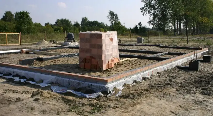 Budowa domu w toku. Widać zarys fundamentów, zbrojenie i ułożoną cegłę. Na środku placu budowy stoi paleta cegieł.