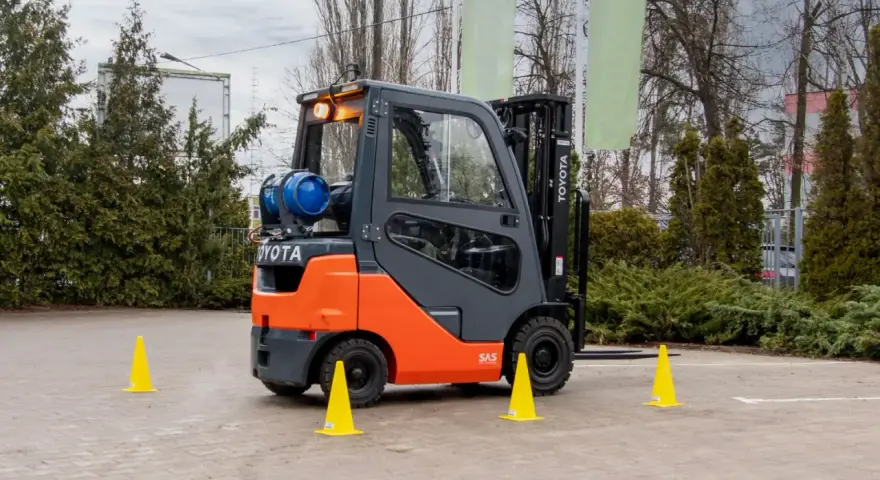 Pomarańczowo-szary wózek widłowy Toyota z butlą gazową, otoczony żółtymi pachołkami. Zastanawiasz się, ile trwa kurs na wózek widłowy?