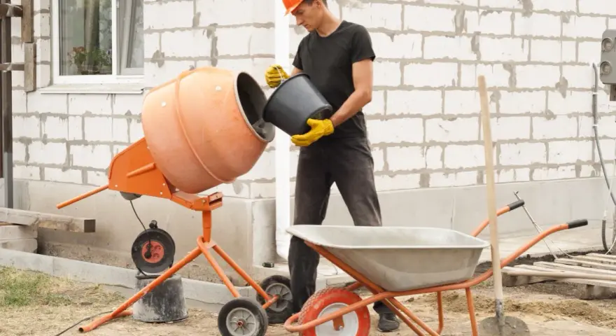 Budowlaniec wsypuje cement do betoniarki 100l. Obok stoi taczka i łopata.