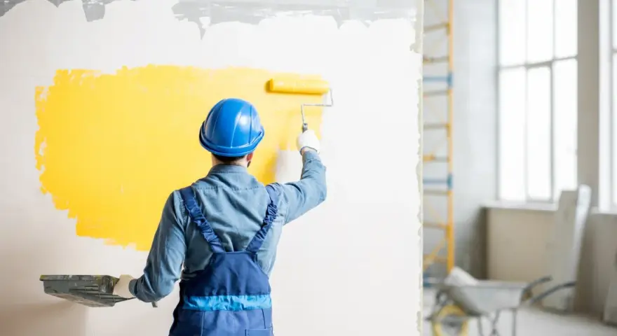 Malowanie ściany na żółto przez pracownika w niebieskim kasku i kombinezonie.