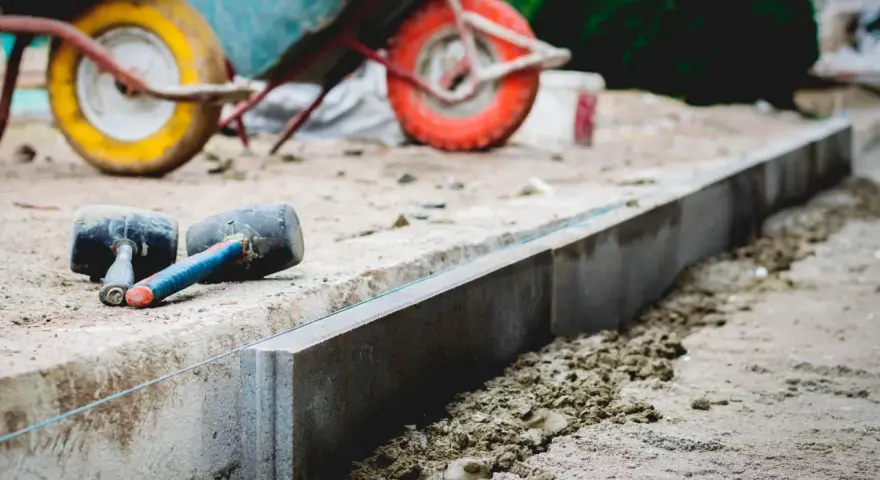 Dwa gumowe młotki leżą obok świeżo ułożonych krawężników. W tle widać taczkę, sugerującą prace budowlane i potrzebę odpowiedniej podsypki pod krawężniki proporcje.
