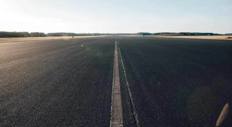 Asfaltowa droga z linią pośrodku, otoczona polami i lasem.