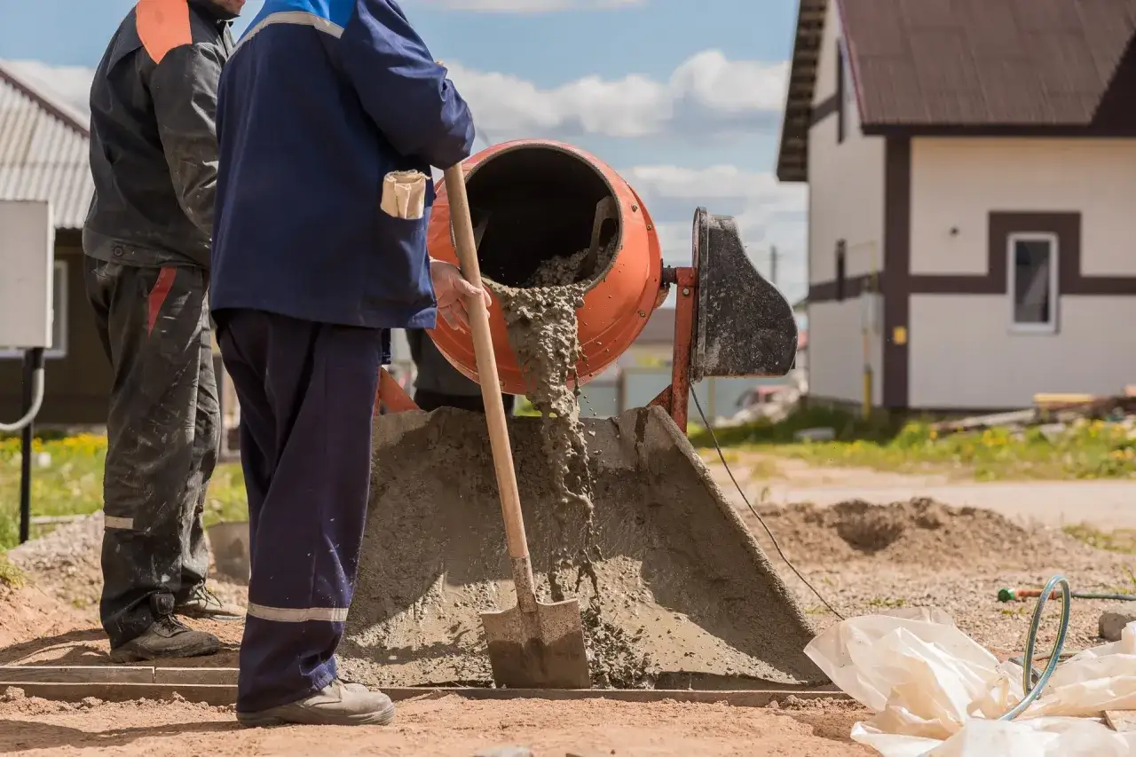 Dwóch budowlańców pracuje przy betoniarce. Jeden z nich miesza łopatą beton, który wylewa się z bębna. Trudno określić, ile łopat żwiru na worek cementu, ale praca wre.