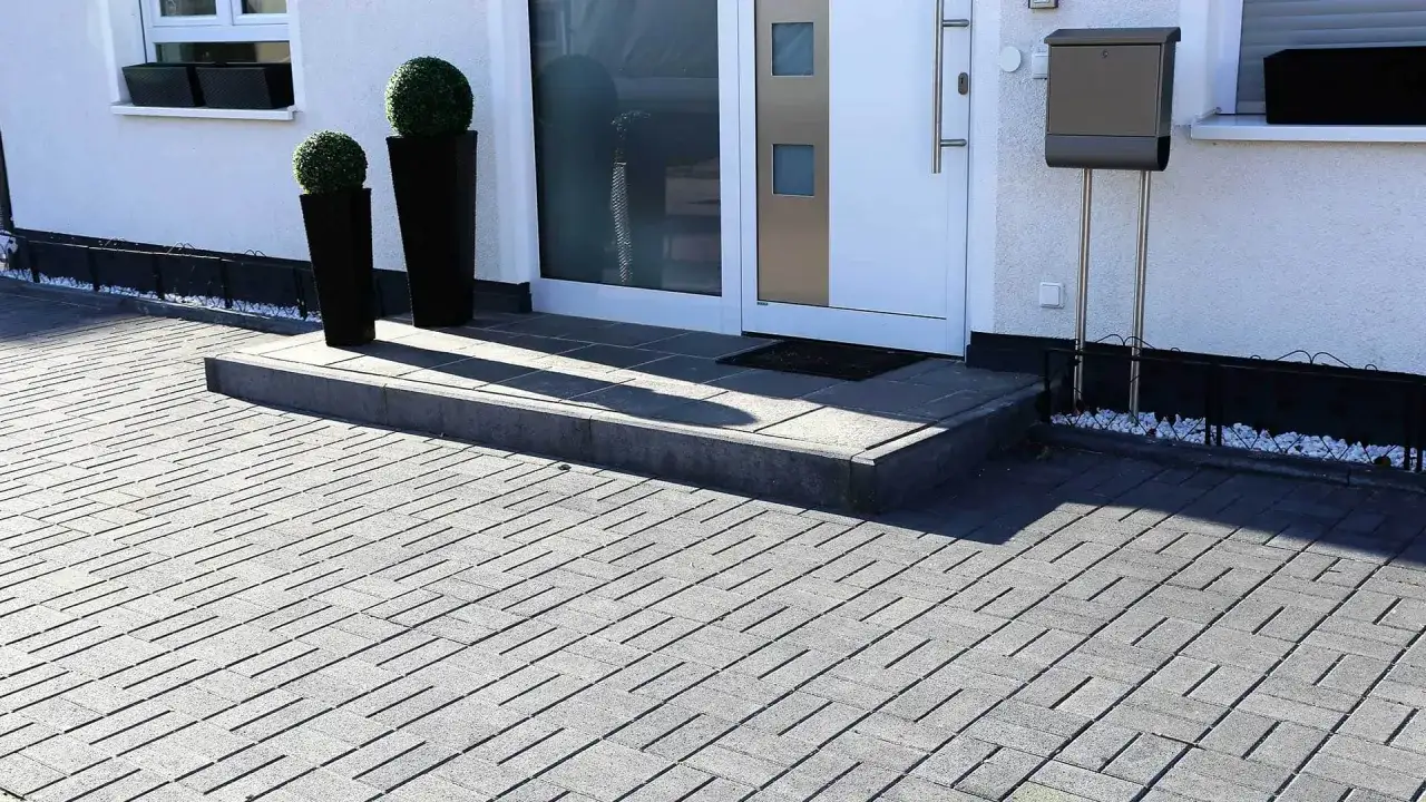 Nowoczesna kostka przed domem tworzy schludny podjazd. Dwa ozdobne krzewy w czarnych donicach witają gości.