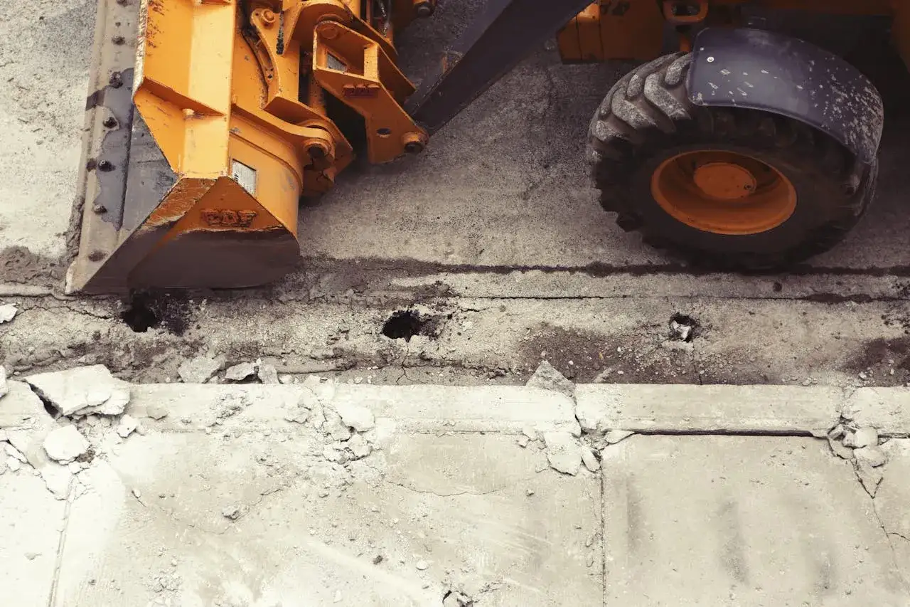 Koparka przygotowuje teren pod wymianę gruntu pod płytę fundamentową. Widać jej żółte ramię i oponę.