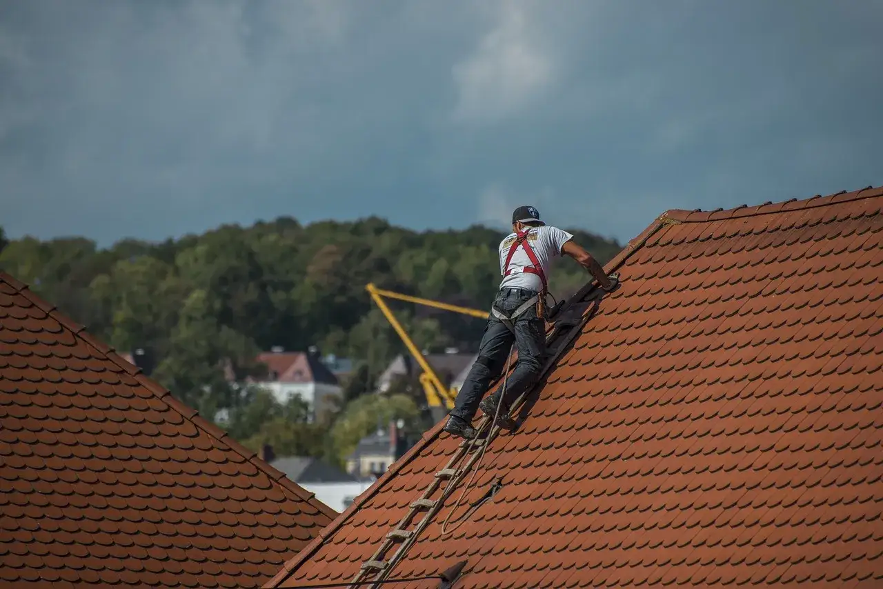 Kto to dekarz? Pracownik na dachu, zabezpieczony linami, układa dachówki. W tle drzewa i budynki.