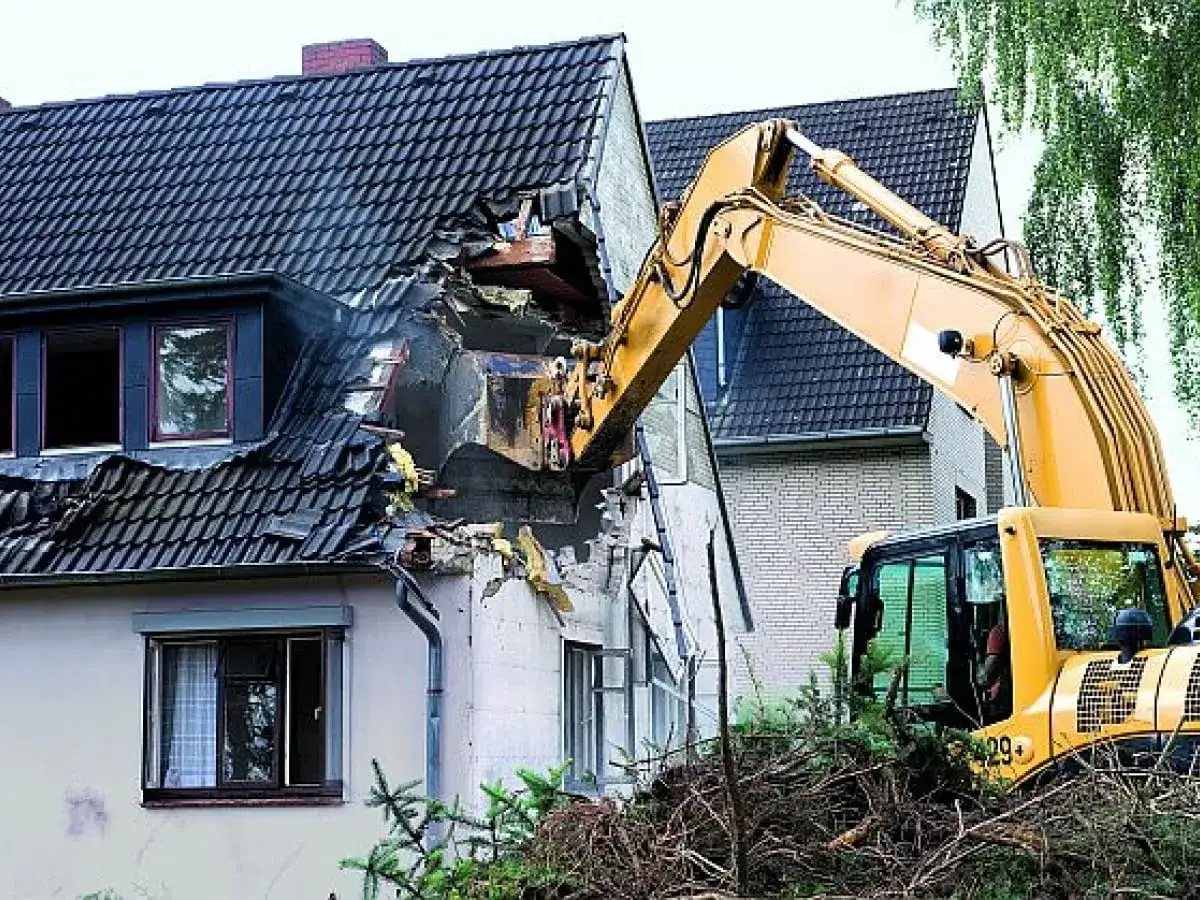 Koparka niszczy dom, pokazując wysoki koszt rozbiórki. Ruiny i gruzy świadczą o postępującym demontażu.