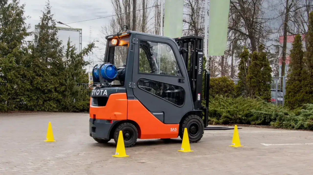 Pomarańczowo-szary wózek widłowy Toyota z butlą gazową, otoczony żółtymi pachołkami. Zastanawiasz się, ile trwa kurs na wózek widłowy?