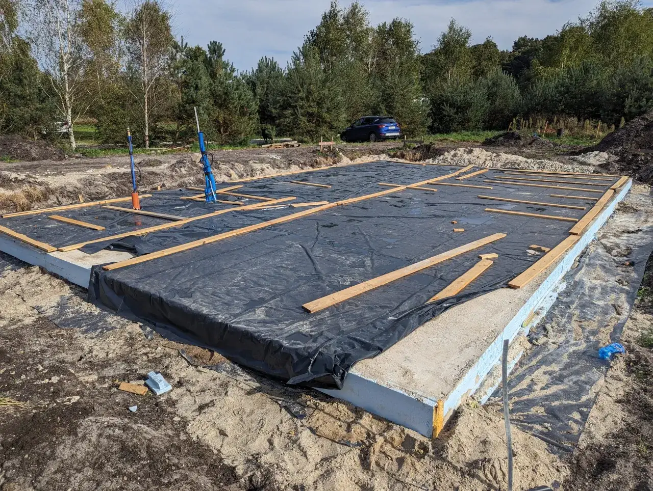 Fundamenty domu w trakcie budowy, przykryte czarną folią i drewnianymi belkami. Czas oczekiwania, ile muszą odstać fundamenty, jest kluczowy dla trwałości konstrukcji.