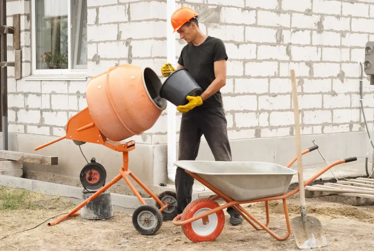 Budowlaniec wsypuje cement do betoniarki 100l. Obok stoi taczka i łopata.
